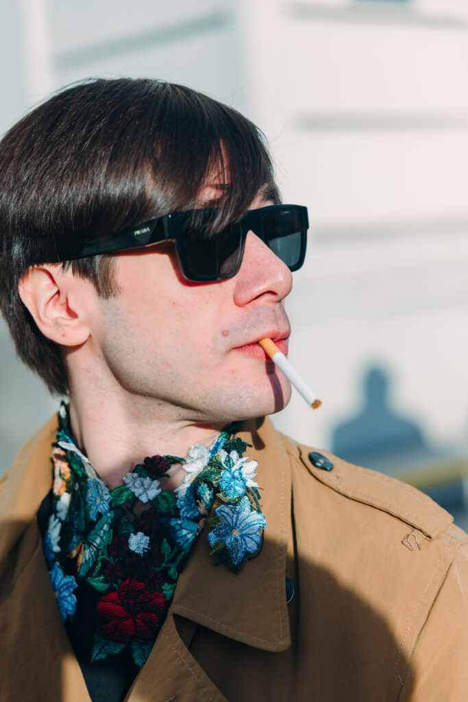 A close-up outdoor portrait of a man wearing black rectangular sunglasses, a beige trench coat, and an embroidered floral collar in vivid red, blue, white, and green tones. He is looking to the side with a cigarette between his lips.